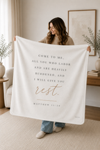 A woman holds up a large cream-colored blanket in a bright, modern living room. The blanket features a printed Bible verse from Matthew 11:28 in dark gray text with 'rest.