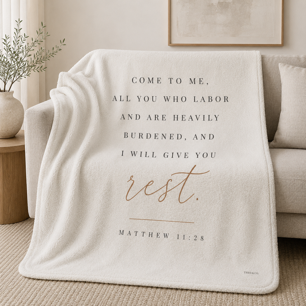 An off-white fleece throw blanket draped over a beige sofa in a softly styled living room. The blanket features printed text reading 'COME TO ME, ALL YOU WHO LABOR AND ARE HEAVILY BURDENED, AND I WILL GIVE YOU rest.