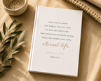 A white hardcover Bible book with elegant black and rose gold typography, styled in a flat-lay arrangement on a beige linen surface with a small ceramic bowl, olive branches, and a woven textile.