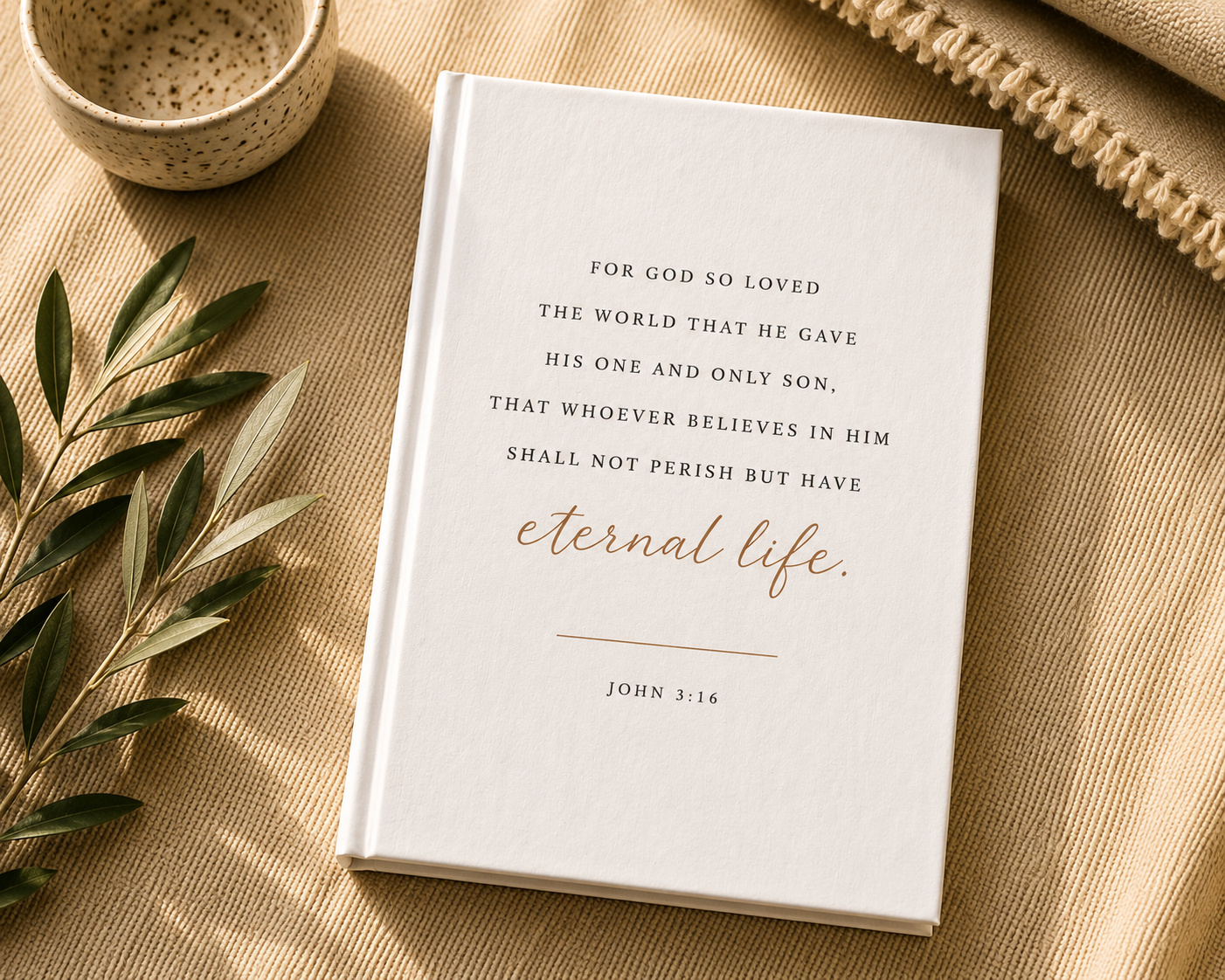 A white hardcover Bible book with elegant black and rose gold typography, styled in a flat-lay arrangement on a beige linen surface with a small ceramic bowl, olive branches, and a woven textile.