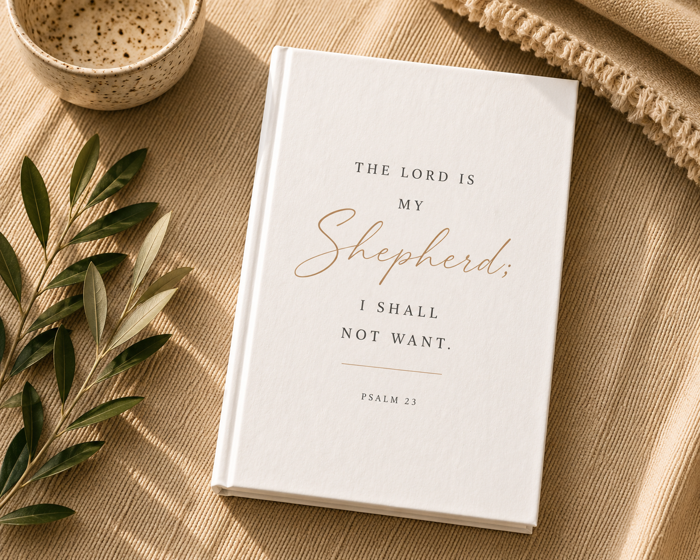 A white hardcover book with the Bible verse 'THE LORD IS MY Shepherd; I SHALL NOT WANT. PSALM 23' printed on the cover, styled in a flat lay on a beige textured fabric with olive branches, a ceramic bowl, and a woven textile.