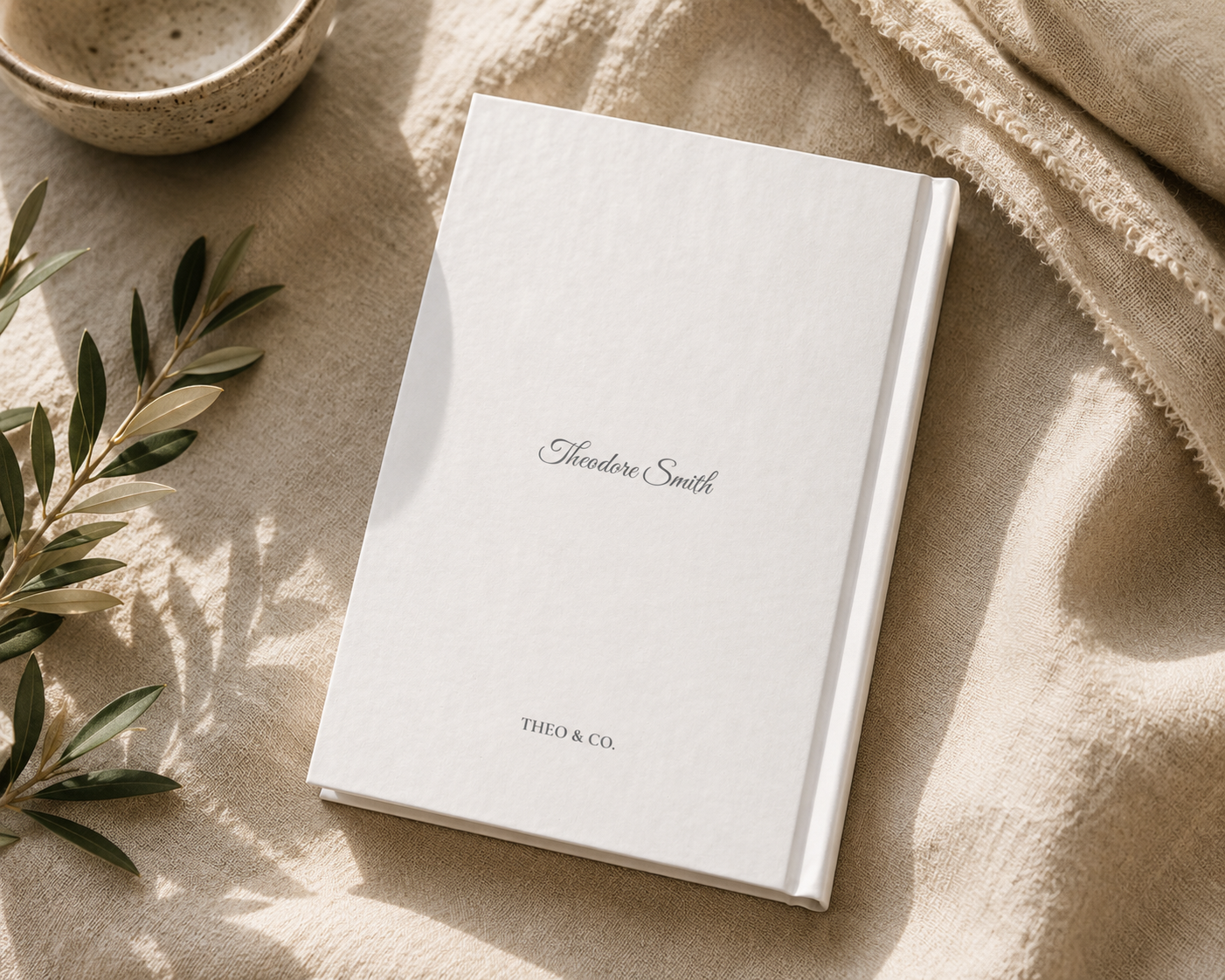A white hardcover notebook or journal with the name 'Alexander Smith' and 'THIO & CO.' printed on the cover, styled on a beige linen surface with a ceramic bowl and green leaves nearby.