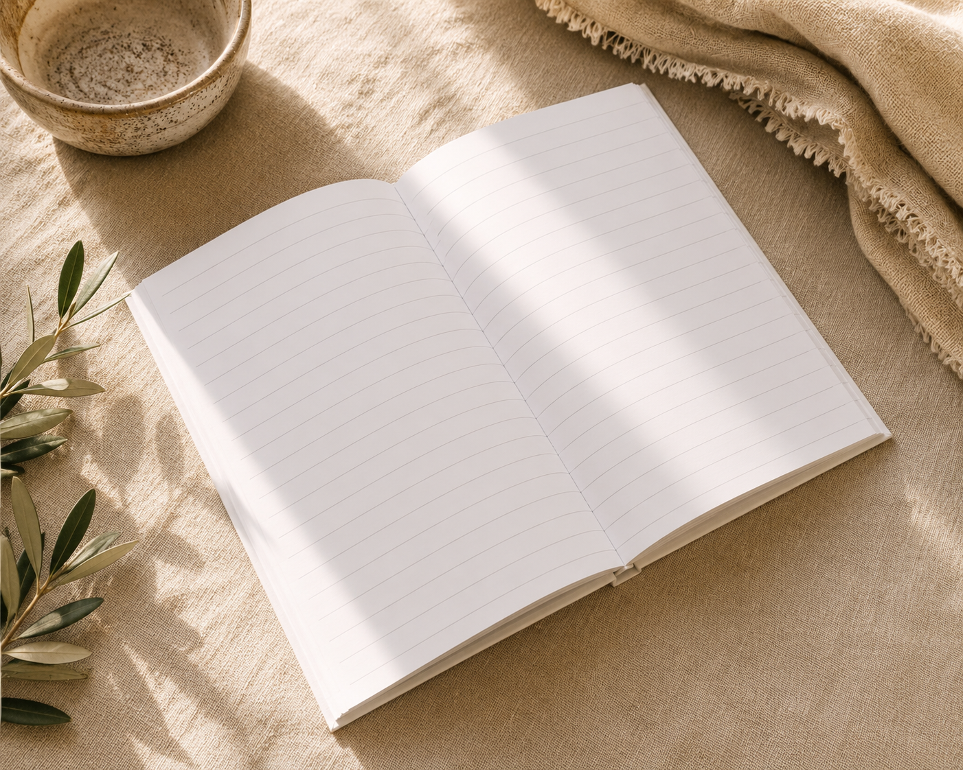 An open lined notebook or journal lies on a beige fabric surface with soft sunlight across the pages. A small ceramic bowl, olive branches, and a folded cream textile are arranged around it in a styled flat-lay setting.