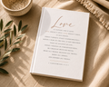 A white hardcover book titled 'Love' with elegant gold script and black scripture text, styled on a beige linen surface with a woven bowl, olive branch leaves, and a fringed textile.