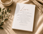 A white hardcover book titled 'Love' with elegant gold script and black scripture text, styled on a beige linen surface with a woven bowl, olive branch leaves, and a fringed textile.