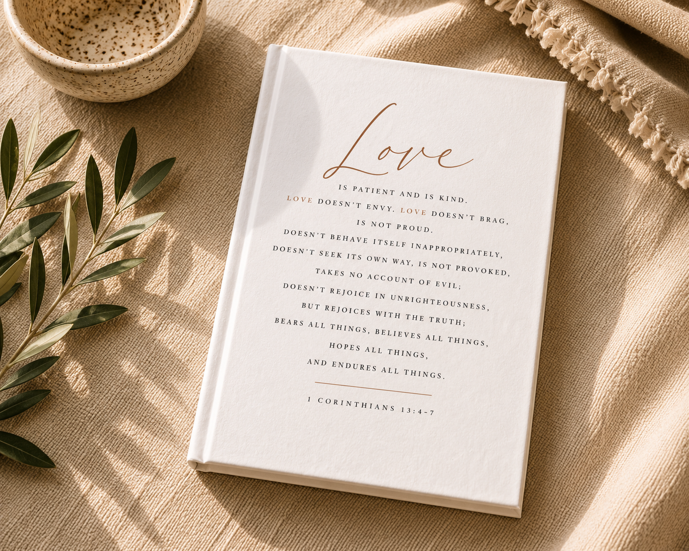 A white hardcover book titled 'Love' with elegant gold script and black scripture text, styled on a beige linen surface with a woven bowl, olive branch leaves, and a fringed textile.