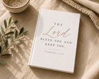 A white hardcover book with elegant rose gold and black typography reading 'THE Lord BLESS YOU AND KEEP YOU. NUMBERS 6:24' lies on a beige linen surface with styled props including a small bowl, olive branch, and textured fabric.