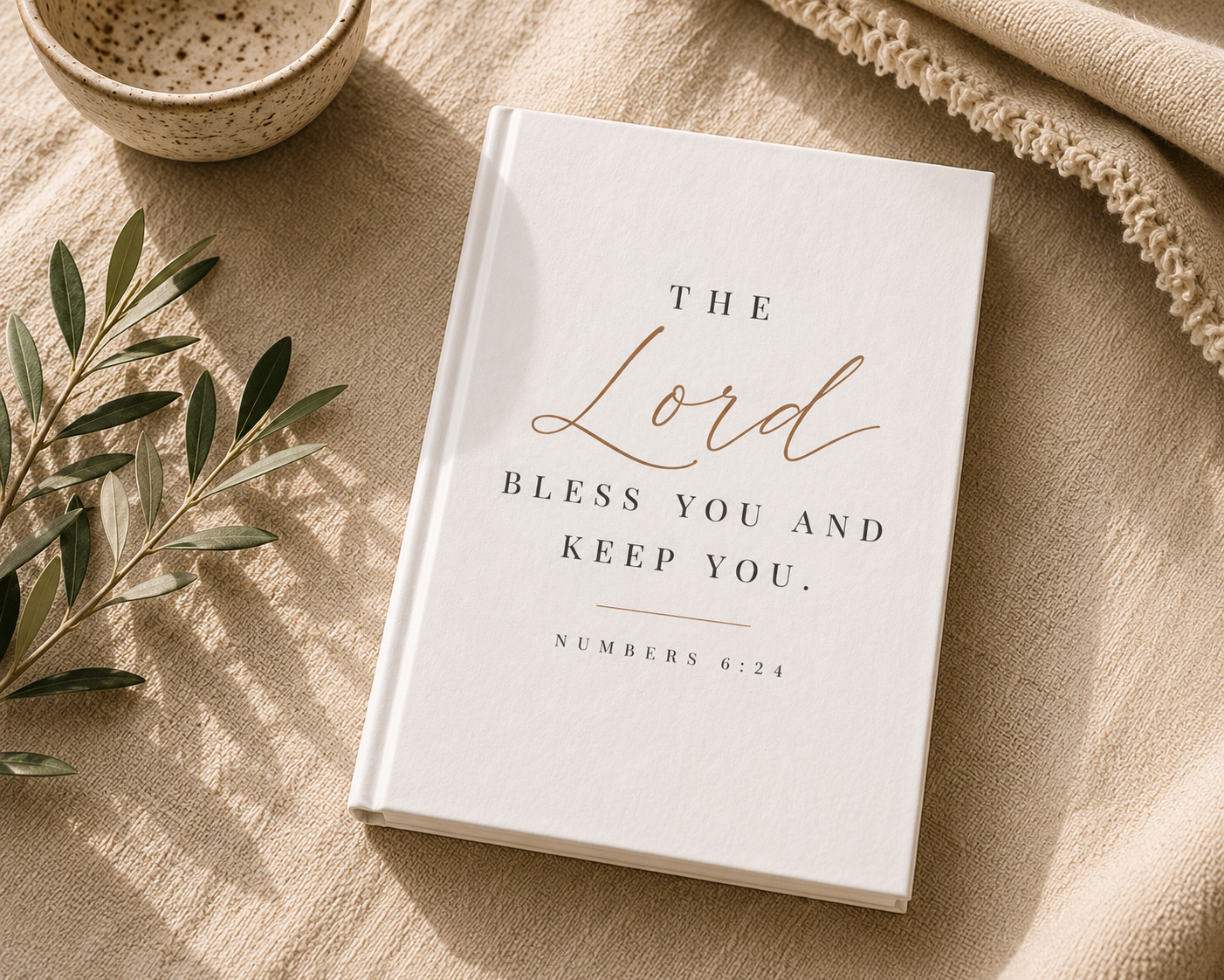 A white hardcover book with elegant rose gold and black typography reading 'THE Lord BLESS YOU AND KEEP YOU. NUMBERS 6:24' lies on a beige linen surface with styled props including a small bowl, olive branch, and textured fabric.
