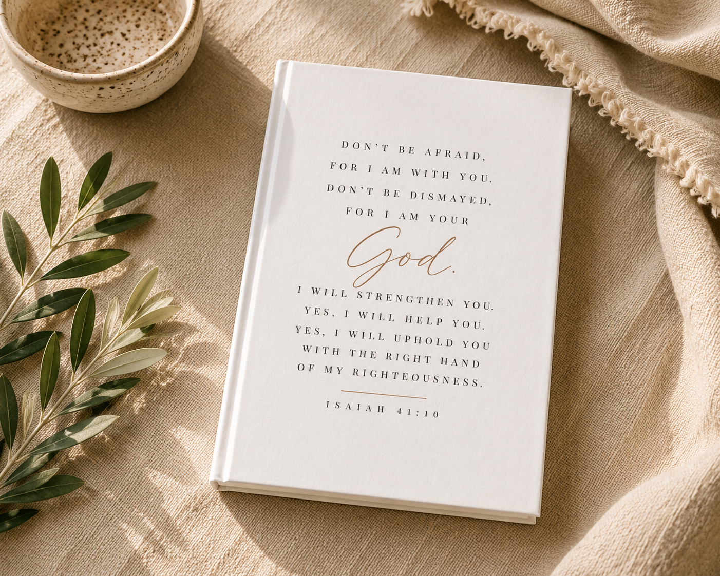A white hardcover journal or Bible-style book with a printed scripture quote in black and rose-gold lettering, styled on a beige linen surface with a small bowl, greenery, and draped fabric.