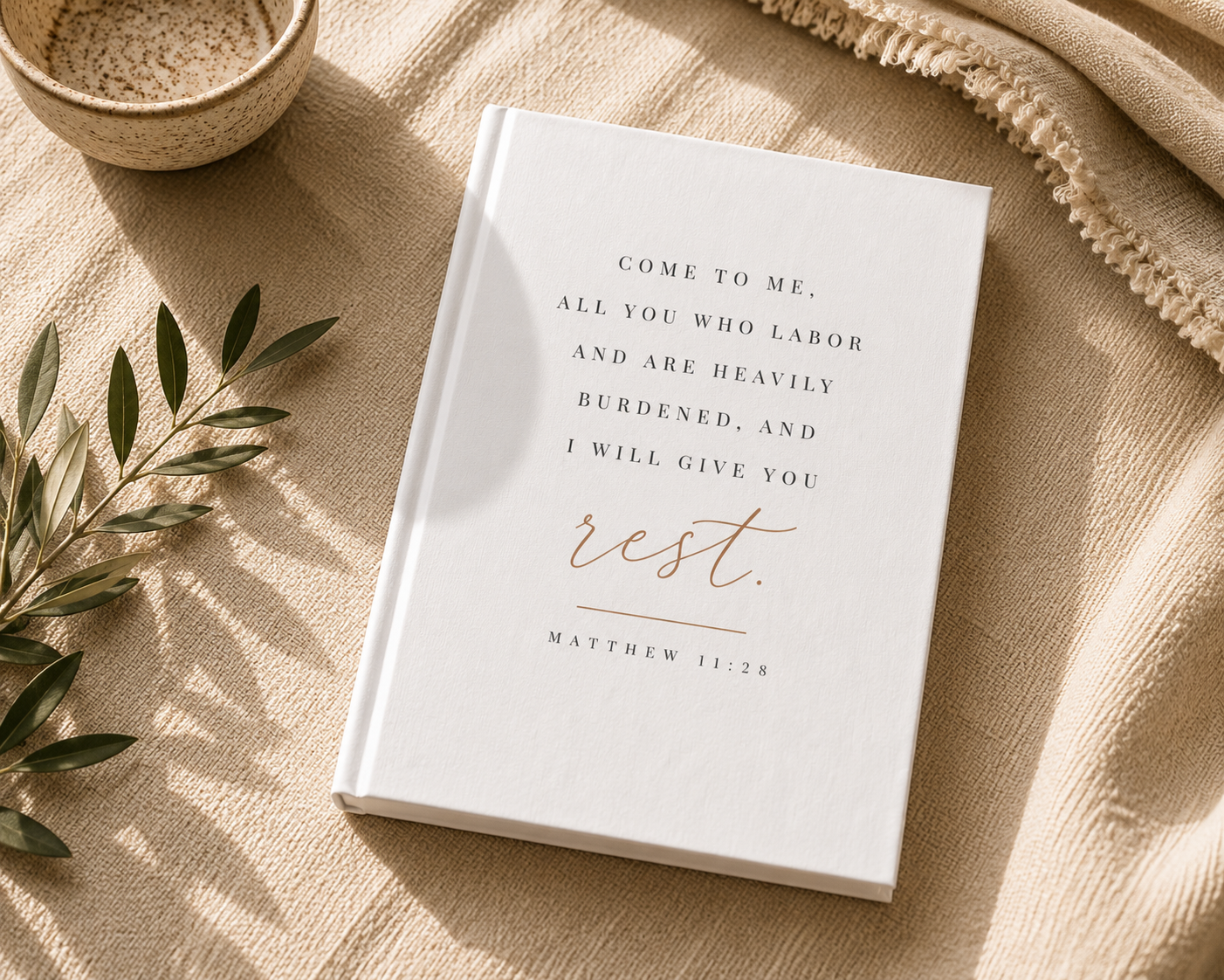 A white hardcover journal or Bible-style book with elegant black and rose gold typography reading 'COME TO ME, ALL YOU WHO LABOR AND ARE HEAVILY BURDENED, AND I WILL GIVE YOU rest. MATTHEW 11:28', styled on a beige textured blanket with a small bowl, greenery, and a fringed textile.