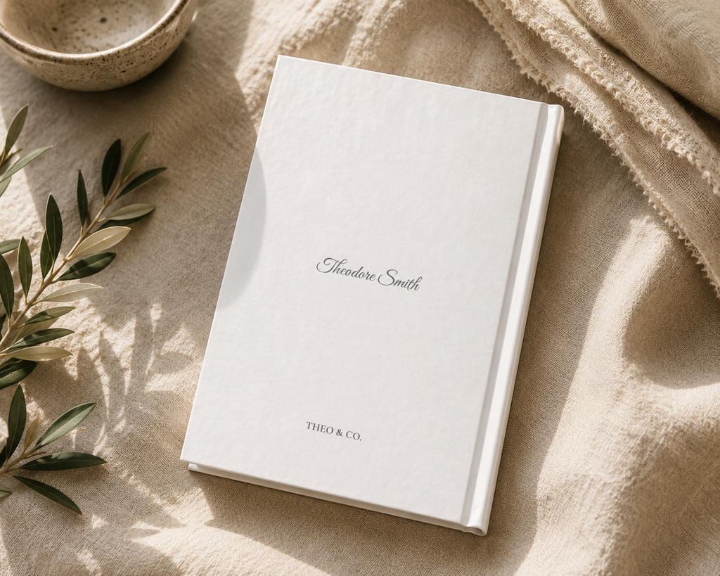 A white hardcover notebook or journal with the name 'Alexander Smith' and 'THIO & CO.' printed on the cover, styled on a beige linen surface with a ceramic bowl and green leaves nearby.