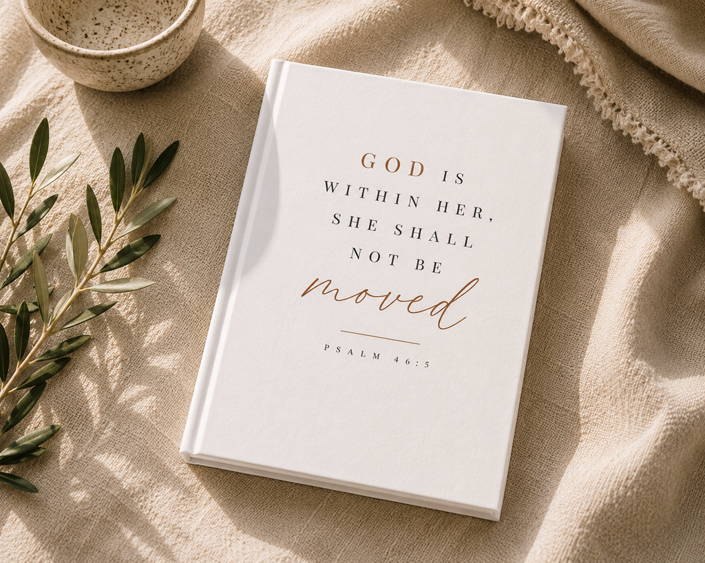 A white hardcover journal or Bible with the printed quote 'GOD IS WITHIN HER, SHE SHALL NOT BE moved' in black and rose-gold lettering, styled on a beige linen fabric with a ceramic bowl, olive branch, and knit blanket.