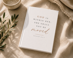 A white hardcover journal or Bible with the printed quote 'GOD IS WITHIN HER, SHE SHALL NOT BE moved' in black and rose-gold lettering, styled on a beige linen fabric with a ceramic bowl, olive branch, and knit blanket.