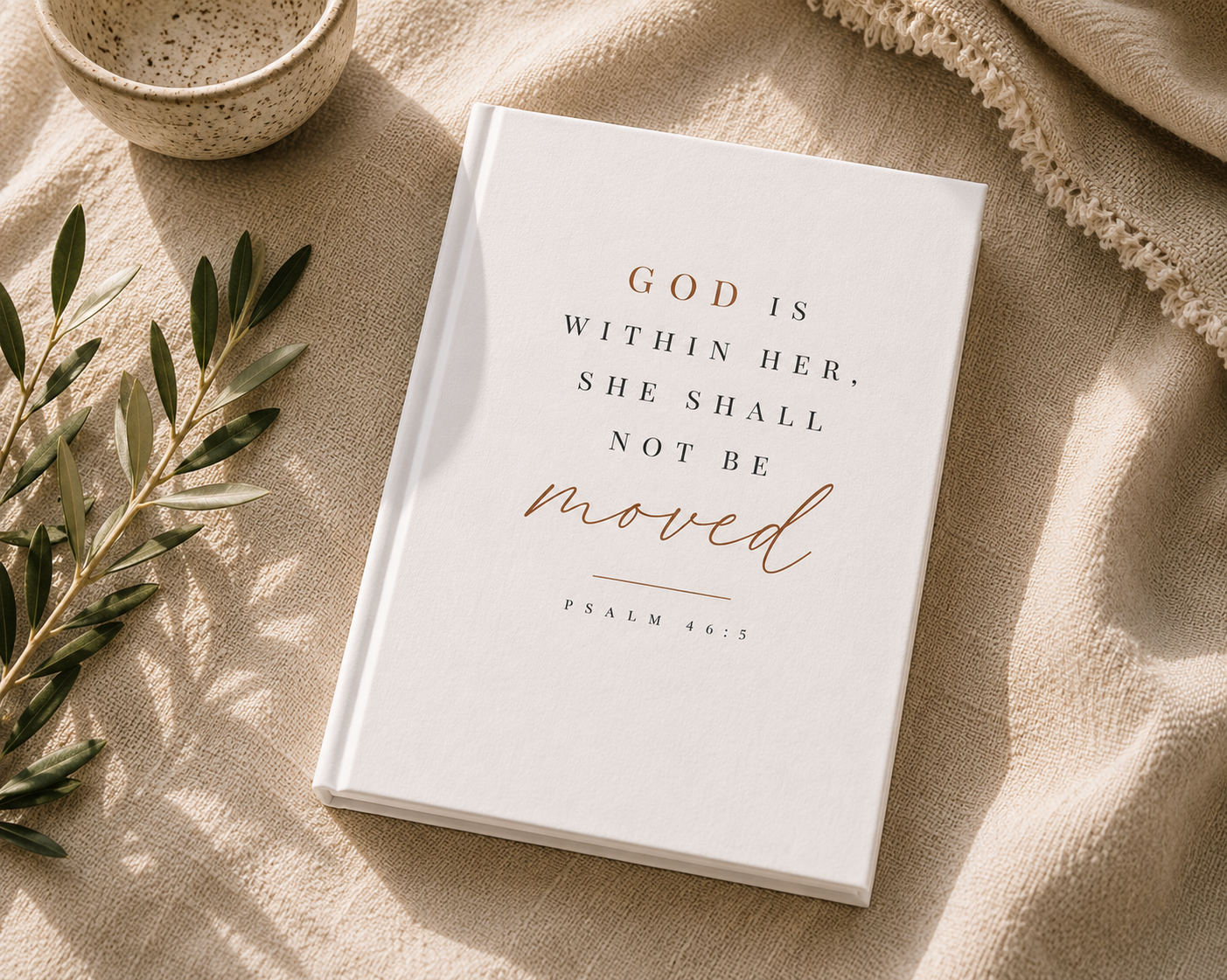 A white hardcover journal or Bible with the printed quote 'GOD IS WITHIN HER, SHE SHALL NOT BE moved' in black and rose-gold lettering, styled on a beige linen fabric with a ceramic bowl, olive branch, and knit blanket.