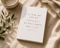 A hardcover book with a white cover and elegant typography reading 'AS FOR ME AND MY house WE WILL SERVE THE LORD' with 'house' in cursive and the verse 'JONAH 24:15' below, styled on a beige linen surface with a ceramic bowl, olive branch, and fringed textile nearby.