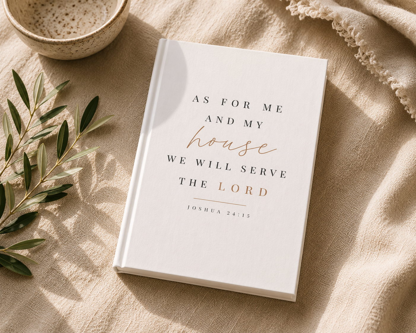 A hardcover book with a white cover and elegant typography reading 'AS FOR ME AND MY house WE WILL SERVE THE LORD' with 'house' in cursive and the verse 'JONAH 24:15' below, styled on a beige linen surface with a ceramic bowl, olive branch, and fringed textile nearby.