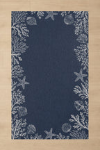 A rectangular navy blue rug with a white nautical border featuring starfish, seashells, and coral, shown flat on a light wood floor.