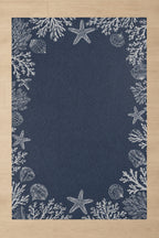 A rectangular navy blue rug with a white seashore print featuring starfish, shells, and coral, laid flat on a light wood floor.