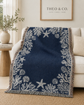 A navy blue throw blanket with a white nautical print of starfish, seashells, and coral, draped over a cream sofa in a styled living room setting.