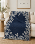A navy blue throw blanket with a white nautical print of starfish, seashells, and coral, draped over a cream sofa in a styled living room setting.