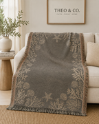A gray throw blanket with beige botanical and starfish patterns draped over a cream sofa in a living room setting.