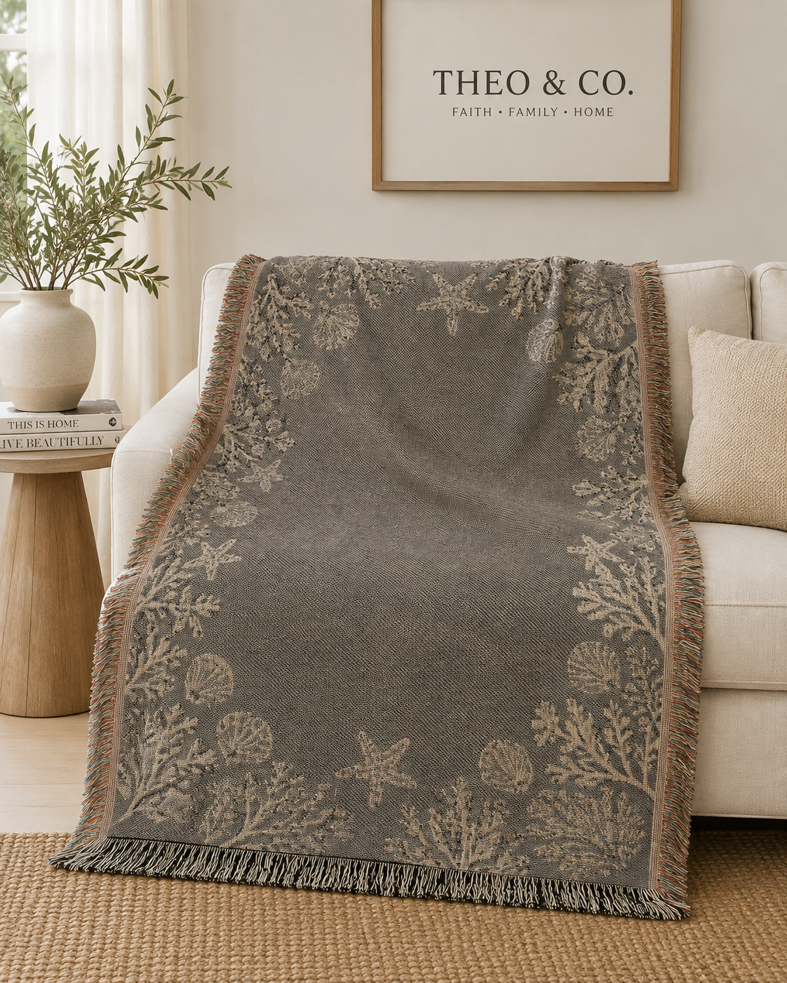 A gray throw blanket with beige botanical and starfish patterns draped over a cream sofa in a living room setting.