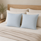 Two square light blue throw pillows displayed on a bed with cream bedding, a wooden headboard, and a neutral-toned side table with a vase.