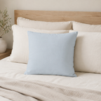 A square light blue throw pillow centered on a bed with cream bedding, white pillows, a wooden headboard, and a beige blanket, with a small plant at the side.
