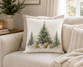 A square decorative throw pillow on a beige sofa, featuring a watercolor-style Christmas tree design with wrapped gifts at the base.