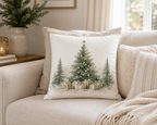 A square decorative throw pillow on a beige sofa, featuring a watercolor-style Christmas tree design with wrapped gifts at the base.