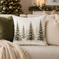A square decorative throw pillow featuring a winter forest design with snow-covered pine trees, styled on a beige sofa with other pillows and a knit blanket, with a decorated Christmas tree in the background.