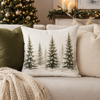 A square decorative throw pillow featuring a winter forest design with snow-covered pine trees, styled on a beige sofa with other pillows and a knit blanket, with a decorated Christmas tree in the background.
