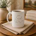 A white ceramic coffee mug with a Bible verse printed on it, sitting on a woven placemat with a folded linen napkin. The mug is filled with a hot beverage that is visibly steaming.