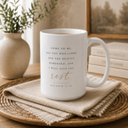 A white ceramic mug with a Bible verse printed on it, placed on a woven placemat with a beige cloth underneath in a cozy home setting.