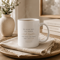 A white ceramic mug with a spiritual quote printed in black and rose gold lettering, resting on a textured beige cloth on a wooden tray. The background features a neutral-toned room with a vase of dried stems and stacked books.