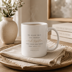 A white ceramic mug with a spiritual quote printed in black and rose gold lettering, resting on a textured beige cloth on a wooden tray. The background features a neutral-toned room with a vase of dried stems and stacked books.