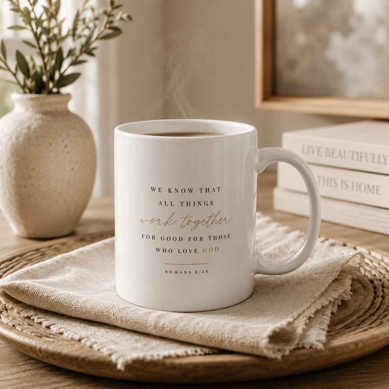 A white ceramic mug with a spiritual quote printed in black and rose gold lettering, resting on a textured beige cloth on a wooden tray. The background features a neutral-toned room with a vase of dried stems and stacked books.