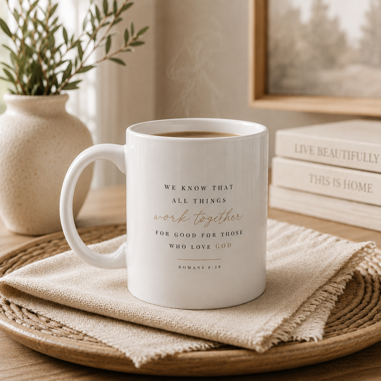 A white ceramic mug with a Bible verse printed on it, sitting on a woven tray with a beige cloth. The background features a potted plant, a stack of books, and soft, warm lighting.