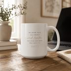 A white ceramic coffee mug with a Bible verse printed on it, sitting on a wooden desk in a cozy, softly lit room with a laptop, notebook, and potted plant in the background.