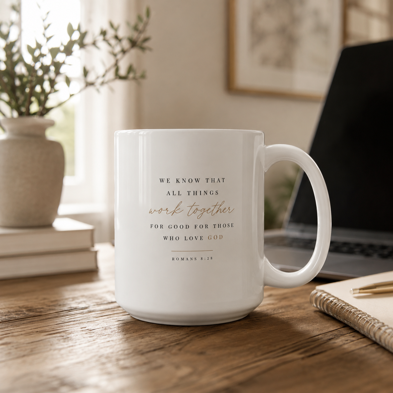 A white ceramic coffee mug with a Bible verse printed on it, sitting on a wooden desk in a cozy, softly lit room with a laptop, notebook, and potted plant in the background.