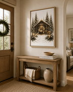 A framed winter forest art print hangs on a beige wall above a rustic wooden console table in a warm, styled entryway. The print depicts a snowy cabin scene with pine trees and warm lantern light.