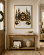 A framed art print depicting a rustic nativity scene with a stable, pine trees, and warm glowing lights, displayed on a beige wall above a wooden console table in a cozy living room setting.