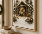 Framed wall art showing a snowy Christmas nativity scene with Mary, Joseph, baby Jesus in a manger, evergreen trees, and a cow, displayed on a beige wall above a wooden surface with books and a bowl.