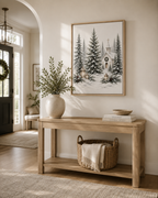 A framed art print depicting a snowy winter forest with pine trees and a small church, styled on a light wood console table in a warm, sunlit entryway.