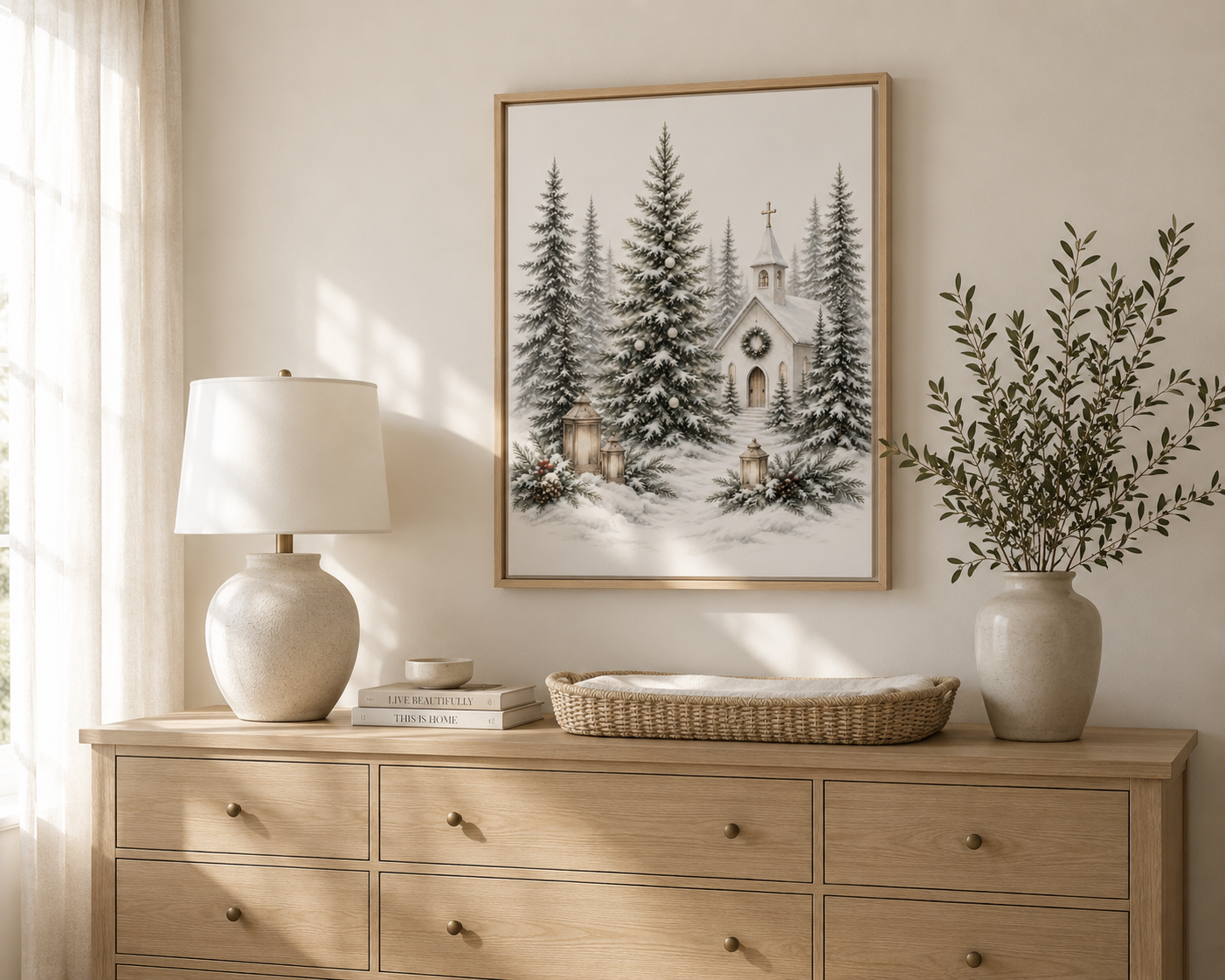 A framed art print depicting a snowy winter scene with pine trees and a small church, styled on a light wood dresser in a bright, neutral-toned room with a lamp and vase.