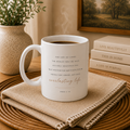 A white ceramic coffee mug with a Bible verse printed on it, sitting on a woven placemat with a burlap napkin. The mug is styled in a cozy home setting with a potted plant, books, and a framed picture in the background.