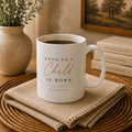 A white ceramic coffee mug with the text 'UNTO USA Child IS BORN ISAIAH 9:6' in black and gold lettering, filled with steaming coffee and placed on a woven placemat with a beige cloth underneath, in a warm home setting.