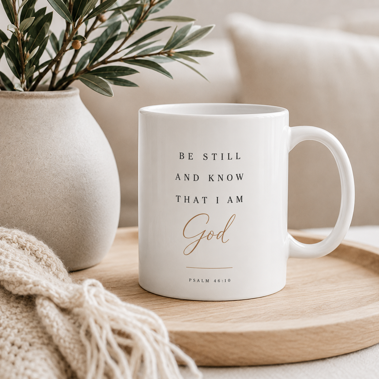 A white ceramic mug with black and gold text reading 'BE STILL AND KNOW THAT I AM God' and 'PSALM 46:10', placed on a round wooden tray beside a beige vase with greenery and a cream knit blanket.