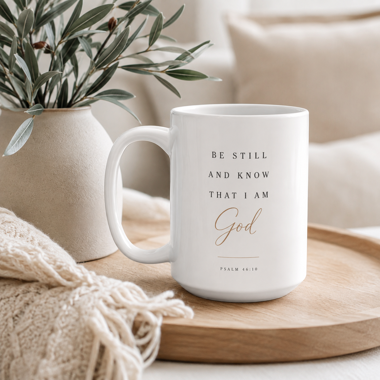 A white ceramic coffee mug with the text 'BE STILL AND KNOW THAT I AM God' in black and rose gold lettering, resting on a wooden tray next to a beige throw blanket and a vase of green leaves.