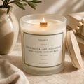 A lit candle in a frosted glass jar with a wooden wick and a printed label reading 'THEO & CO.' and a Bible verse, shown on a beige surface with a wooden lid beside it and soft decorative props in the background.